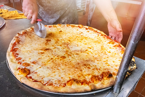Pizzaiolo in New York City cutting a giant cheese pizza at a pizzeria kitchen to take out during Covid-19 state of emergency