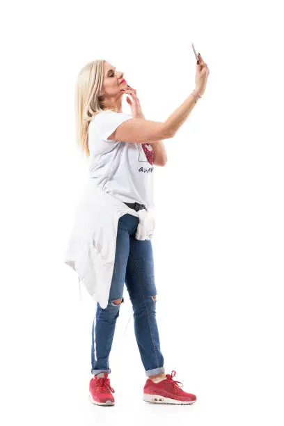 Side view of mid age blonde modern woman in casual clothes taking selfie with hand on chin. Full body length isolated on white background.