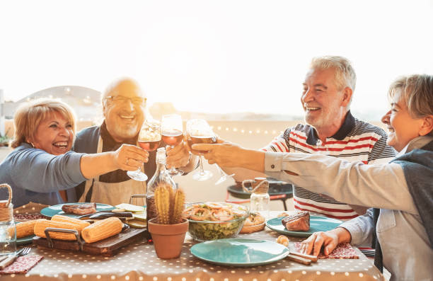 restauration senior heureuse et verres de vin rouge de dégustation dans le dîner de barbecue - famille ayant l’amusement appréciant le bbq à l’heure de coucher du coucher du soleil sur la terrasse - le mode de vie des personnes âgées et le concep - dining senior adult friendship mature adult photos et images de collection