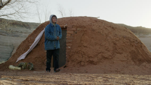 A Native American (Navajo) Teenaged Boy Opens the Door and Exits a Mud Hogan (Navajo Hut) with a Large Rock Wall/Mesa in the Background