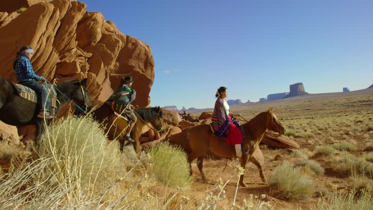 Slow Motion Panning Shot of Several Young Native American (Navajo) Children Wearing Traditional Navajo Clothing and Riding Their Horses through the Landscape of the Monument Valley Desert in Arizona/Utah Next to a Large Rock Formation on a Clear, Bright D