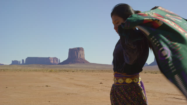 Slow Motion Shot of a Teenaged Native American Girl Wrapping a Traditional Navajo Blanket around Her Shoulders in the Monument Valley Desert with Large Rock Formations in the Distance on a Clear, Bright Day