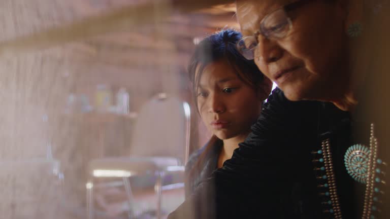 A Native American Grandmother (Navajo) in Her Sixties Teaches Her Teenaged Granddaughter How to Weave at a Loom Indoors