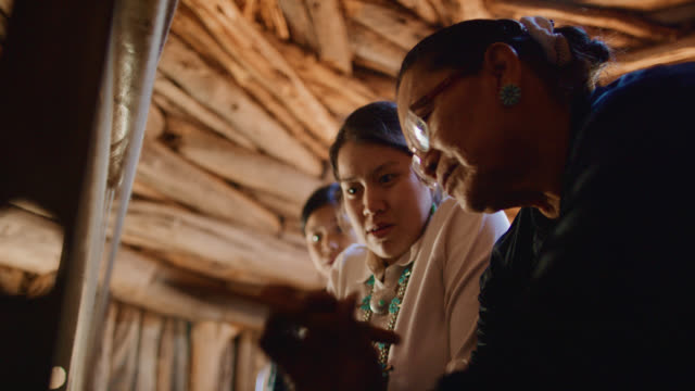 Slow Motion Shot of a Native American Grandmother (Navajo) in Her Sixties Teaching Her Teenaged Granddaughters How to Weave at a Loom Indoors in a Hogan (Navajo Hut)