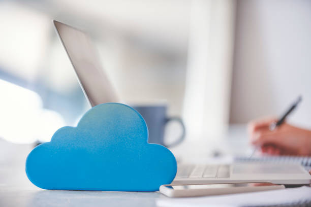 Woman using a laptop with cloud icon on working at home. Woman using a laptop with cloud icon on working at home. She is in the dining room at the dining room table. She is wearing casual clothing. There is a cloud icon next to her computer illustrating a cloud computing network and storage concept. cloud storage stock pictures, royalty-free photos & images