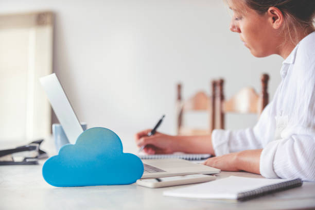 Woman using a laptop with cloud icon on working at home. She is in the dining room at the dining room table. She is wearing casual clothing. There is a cloud icon next to her computer illustrating a cloud computing network and storage concept. mujer usando un ordenador portátil con icono de nube sobre el trabajo en casa. está en el comedor de la mesa del comedor. lleva ropa casual. hay un icono de nube junto a su computadora que ilustra un concepto de almacenamiento y red de computación en la