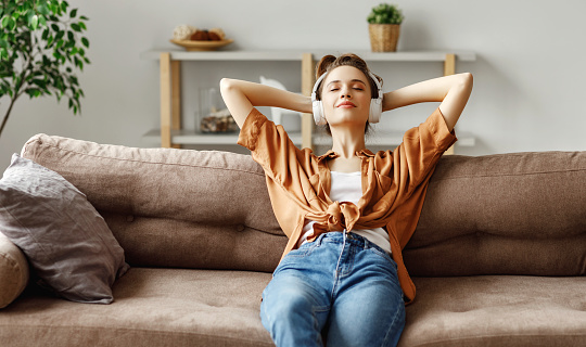 Happy millennial female with eyes closed in wireless headphones and casual clothes sitting alone on soft couch with hands behind head and listening with pleasure to favorite music in light spacious living room