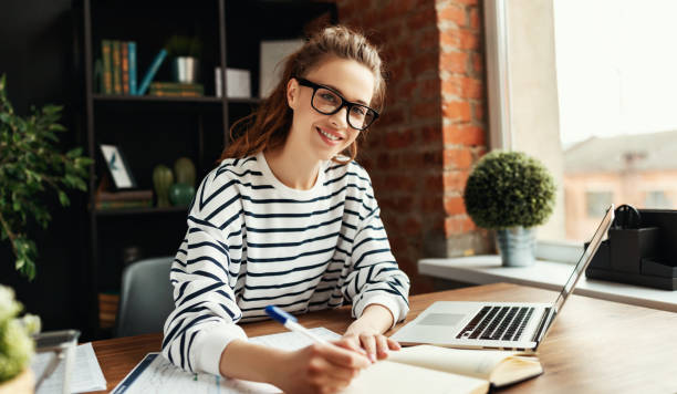 istock Happy millennial woman taking notes in notepad while working at laptop in comfortable loft office 1217255270