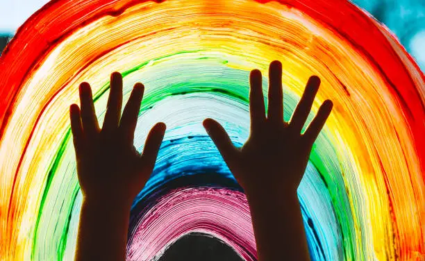 Photo of Close-up photo of child's hands touch painting rainbow on window. Family life background. Image of kids leisure at home, childcare, safety joy symbol.