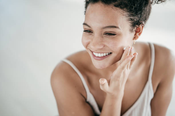 smiling young women applying moisturiser to her face - beauty treatment moisturizer human skin cosmetics imagens e fotografias de stock