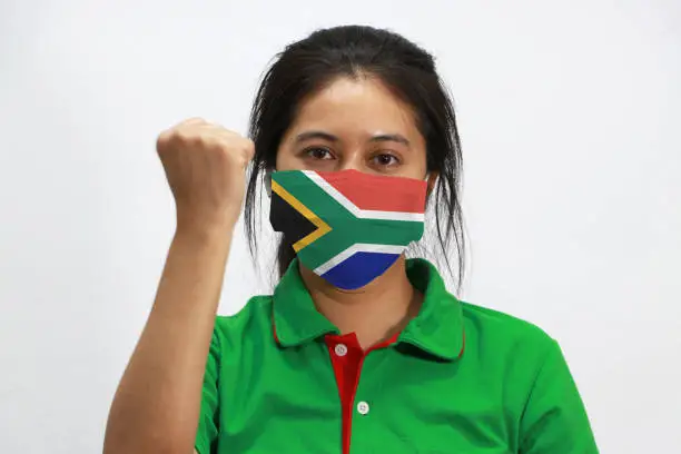 Photo of South Africa flag on hygienic mask. Masked woman prevent germs and wear green shirt. Tiny Particle or virus corona or Covid-19 protection. Lift the fist up for meaning fighting.