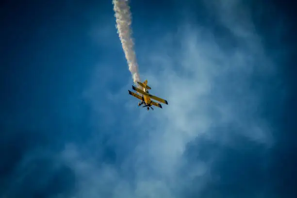 A yellow bi-plane trailing smoke against a blue cloudy sky.