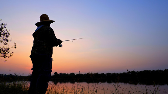 Wide shot of teenage boy standing on the riverbank, holding a fishing rod and enjoying fishing during golden hour sunset or sunrise.