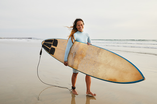 Portrait of a smiling mature woman wearing a wetsuit walking on a beach with her surfboard to the ocean