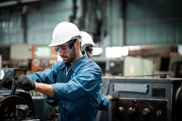 Men industrial engineer wearing a white helmet while standing in a heavy industrial factory behind. The Maintenance looking of working at industrial machinery and check security system in factory. stock photo