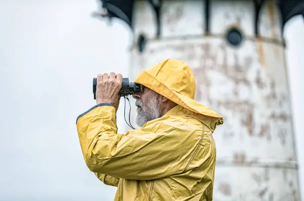 A senior adult ancient mariner gray beard sailor sea boat captain lighthouse keeper man is wearing a bright yellow waterproof fisherman's rain hat and matching rain slicker raincoat rain jacket. He's standing near his lighthouse and looking through magnifying binoculars out over the Pacific Ocean water along the California coastline. Brief code #686998459 - "Senior Men Portraits".