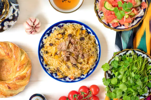 National Uzbek pilaf with meat, achichuk salad of tomato, cucumber, onion in plate with traditional pattern, cilantro, cherry tomatoes, garlicbread tortilla - patir on white wooden table Top view.
