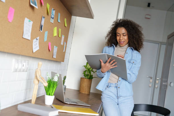 istock Smiling african female college student studying from home, elearning, stand at home office desk, holding notebook making notes. Young mixed race teen girl remote pupil learning doing school homework. 1217312598