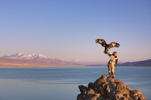 Traditional kazakh eagle hunter with his golden eagle in front of snow capped mountains at a lake shore. Ulgii, Western Mongolia.