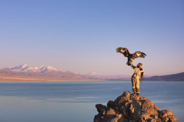 Young kazakh eagle hunter with his golden eagle. Traditional kazakh eagle hunter with his golden eagle in front of snow capped mountains at a lake shore. Ulgii, Western Mongolia. mongolian ethnicity stock pictures, royalty-free photos & images