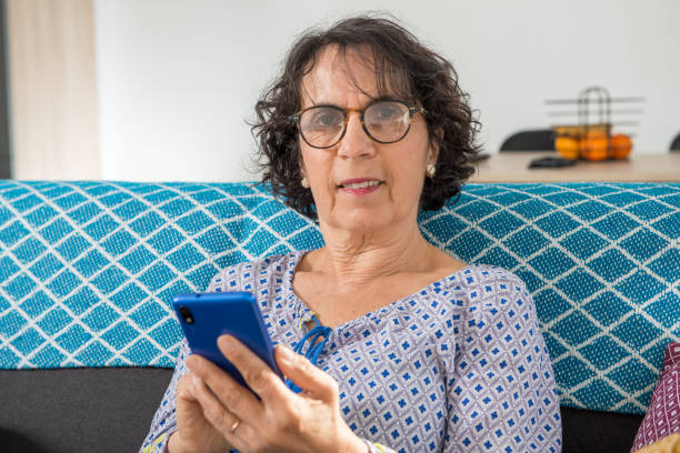 istock cheerful brunette senior woman using smartphone while sitting on sofa 1217316671
