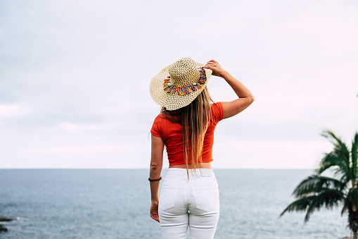 Beautiful unrecognizable young woman viewed from back looking and enjoying the ocean view and feeling - concept of trendy people and outdoor vacation place