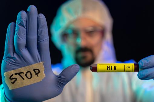 Lab scientist looking at camera and show stop sign while holding HIV (AIDS) blood sample in laboratory. The scene is situated in controlled photography studio environment faking chemical laboratory.  Picture  is taken with Sony A7III camera.
