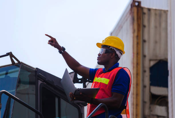 istock Worker man in hardhat and safety vest holding laptop standing on container stackers control loading containers box from cargo 1217329900