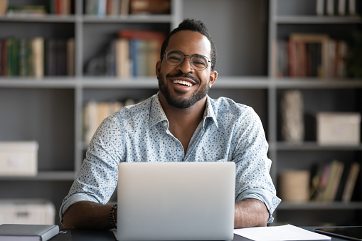Portrait of smiling African American man in glasses sit at desk in office working on laptop, happy biracial male worker look at camera posing, busy using modern computer gadget at workplace