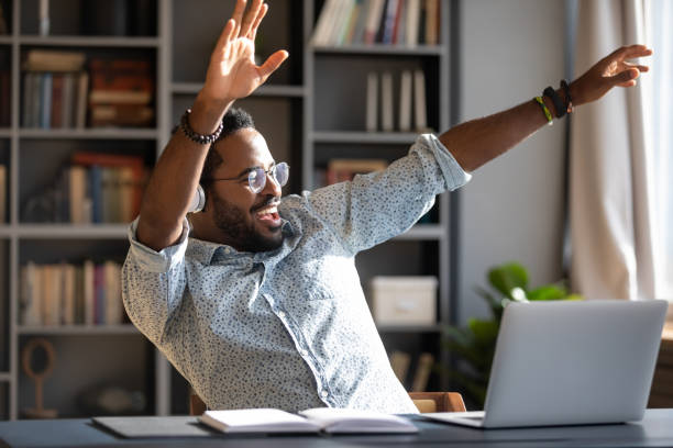 Overjoyed biracial man have fun listening to music Overjoyed young African American man have fun dancing at workplace listening to music in headphones, happy biracial male worker or student enjoy good quality sound in modern wireless earphones excitement laptop stock pictures, royalty-free photos & images