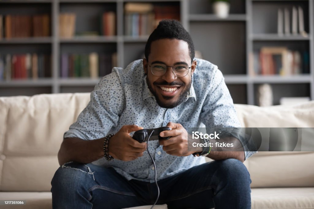 Happy african American man play video game Overjoyed African American millennial man relax on couch in living room play video game using joystick gamepad, happy biracial young male gamer rest at home engaged in digital virtual activity Playing Stock Photo