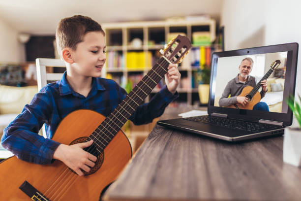 bambino felice che impara a suonare la chitarra mentre guarda le lezioni al laptop a casa - guitar child music learning foto e immagini stock