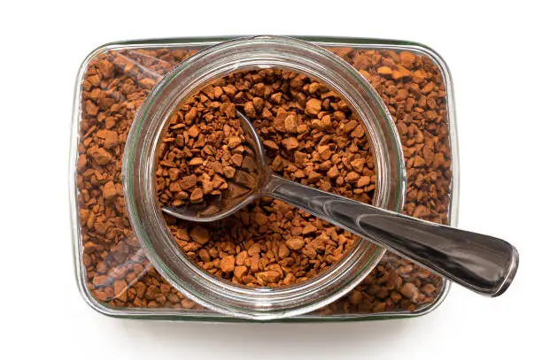 Dry instant coffee in an open glass jar with metal spoon isolated on white. Top view.