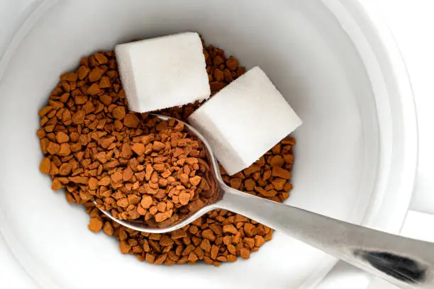 Dry instant coffee and cubes of sugar in a white ceramic cup with metal spoon. Top view.