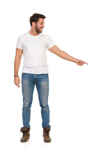 Smiling Handsome Man In Jeans And White T-shirt Is Standing And Pointing Down. Front View.