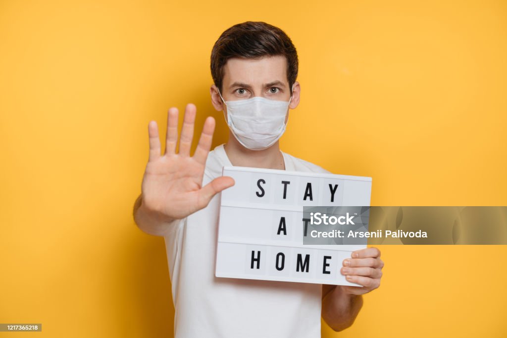 Gue showing stop gesture and stay home text as social media campaign for coronavirus prevention Handsome young man in protective mask holds Stay at home sign and shows stop gesture to prevent coronavirus infection. Yellow background. 
Self isolation and social distancing concept. Viral infection Spreading Stock Photo
