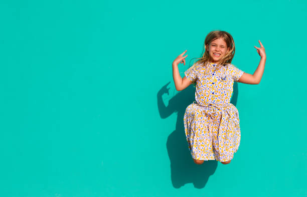 Cheerful little girl in colorful dress showing goat gesture and jumping against green wall in sunlight