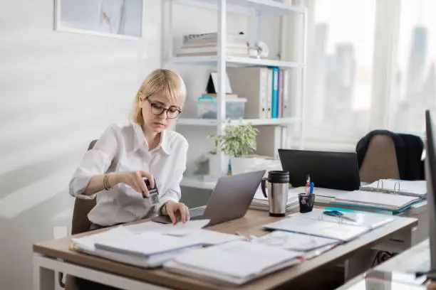 Photo of Young blonde woman with glasses doing paperwork on her desk in a bright office downtown. Junior manager putting a stamp on a document before going to a meeting.