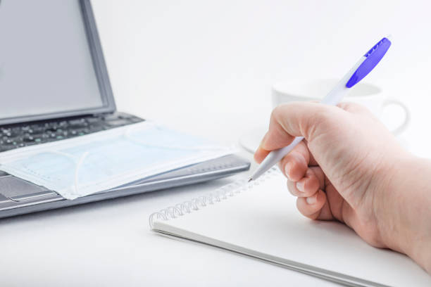 Female hand makes notes in a notebook near a laptop with a medical mask. The concept of working at home business, home schooling, shopping, home purchases. Work during a virus outbreak. Insulation. la mano femenina hace notas en un portátil cerca de un portátil con una máscara médica. el concepto de trabajar en el negocio del hogar, la educación en el hogar, las compras, las compras de casas. trabajar durante un brote de virus. aislamiento.