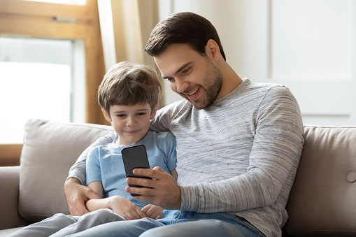 Happy young father sit relax on couch in living room with little son make selfie on smartphone, dad rest on sofa at home with small boy child watch video on cellphone enjoy leisure weekend together