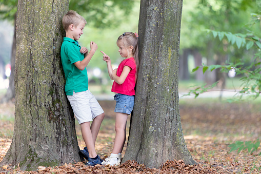 Portrait of two pretty cute children boy and girl standing near big tree trunk in summer park outdoors.