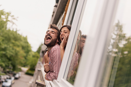 Beautiful young couple leaning through an opened apartment window, waving and shouting, calling friends passing by in the street