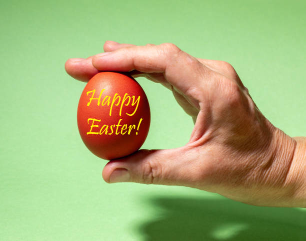 istock Female hand gently holding one traditional colored red Easter egg with inscription Happy Easter. 1217410841