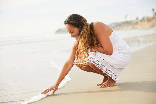 sonriendo mujer madura tocando el oleaje entrante en una playa - women sarong beach white fotografías e imágenes de stock