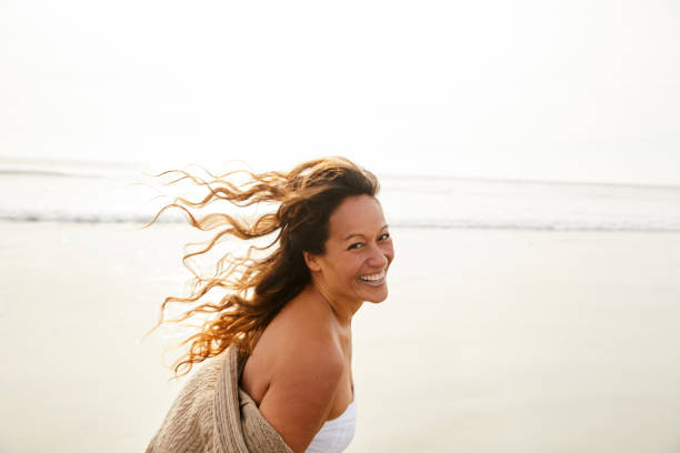 mujer madura riendo caminando en una playa en una tarde ventosa - mature women fotografías e imágenes de stock