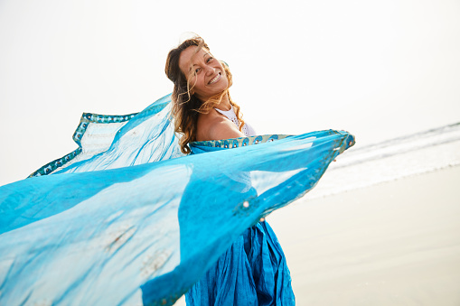 Portrait of a carefree mature woman laughing while dancing with a turquoise wrap along a beach in the late afternoon