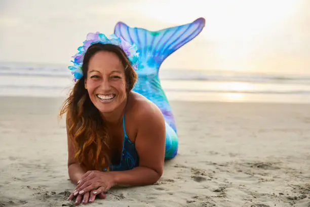 Portrait of a mature woman wearing a mermaid costume laughing while lying on a sandy beach at sunset