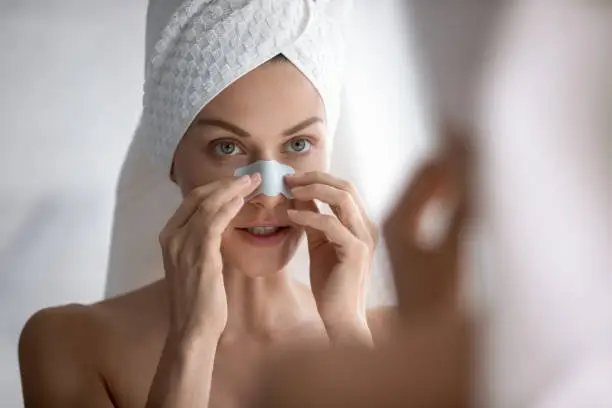 Focused young beautiful woman with towel on head applying cleaning strip, removing dark spots. Attractive lady using nose mask, peeling blackheads blemish, healthy skin care process after showering.