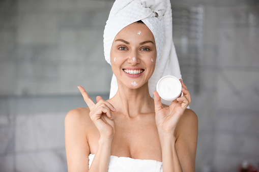 Head shot young smiling woman wrapped in towels holding cream, satisfied with professional cosmetic product results. Happy lady applies make up base on skin after morning skincare routine in bathroom.