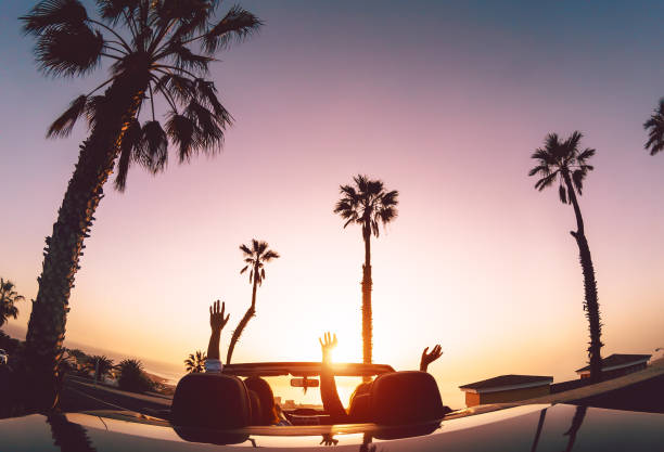 couples heureux ayant l’amusement pendant le voyage de route dans la voiture décapotable - jeunes amants appréciant des vacances dans la ville tropicale - relation d’amour et concept de mode de vie de personnes de voyage - usa travel sunset summer photos et images de collection